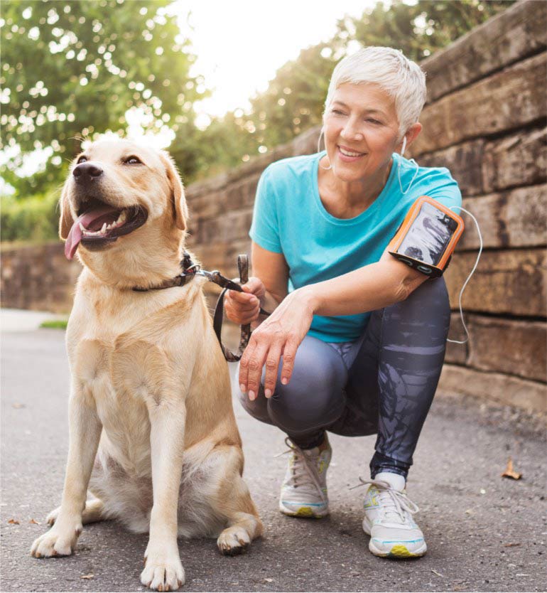 Senior woman in athletic wear kneeling beside happy yellow Labrador outdoors on a sunny day.