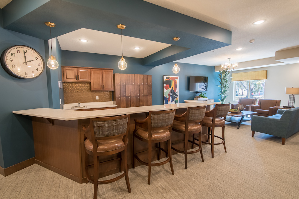 Modern apartment lounge with bar seating, cozy seating area, large clock, and abstract art.