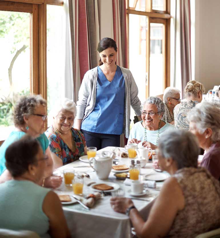 senior-assisted-living-dining Senior women sharing a meal and smiling with a caregiver in the dining area of an assisted living facility.