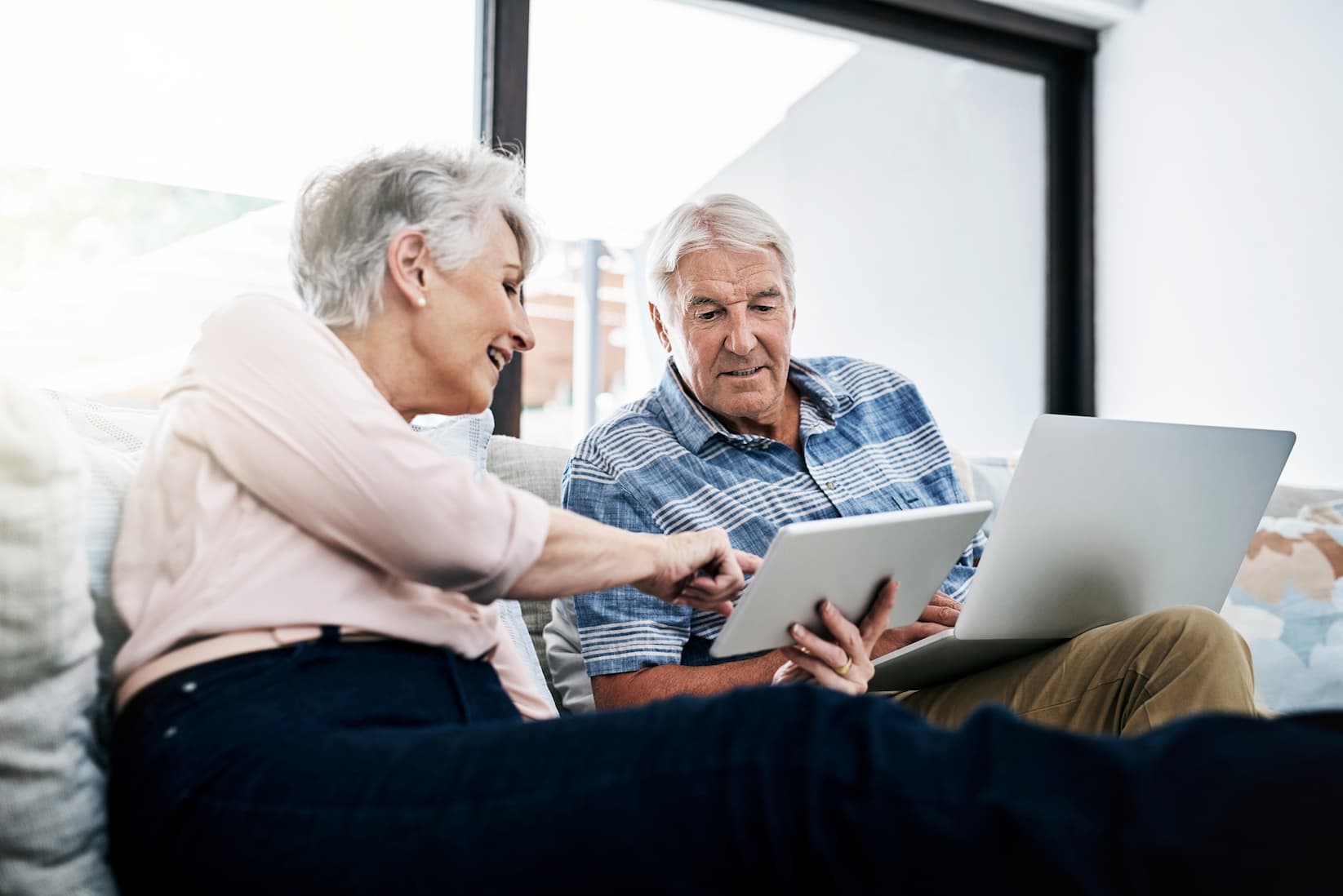 senior-man-and-woman-reviewing-on-laptop Senior man and woman sit on a couch and review information on a laptop and tablet screen together