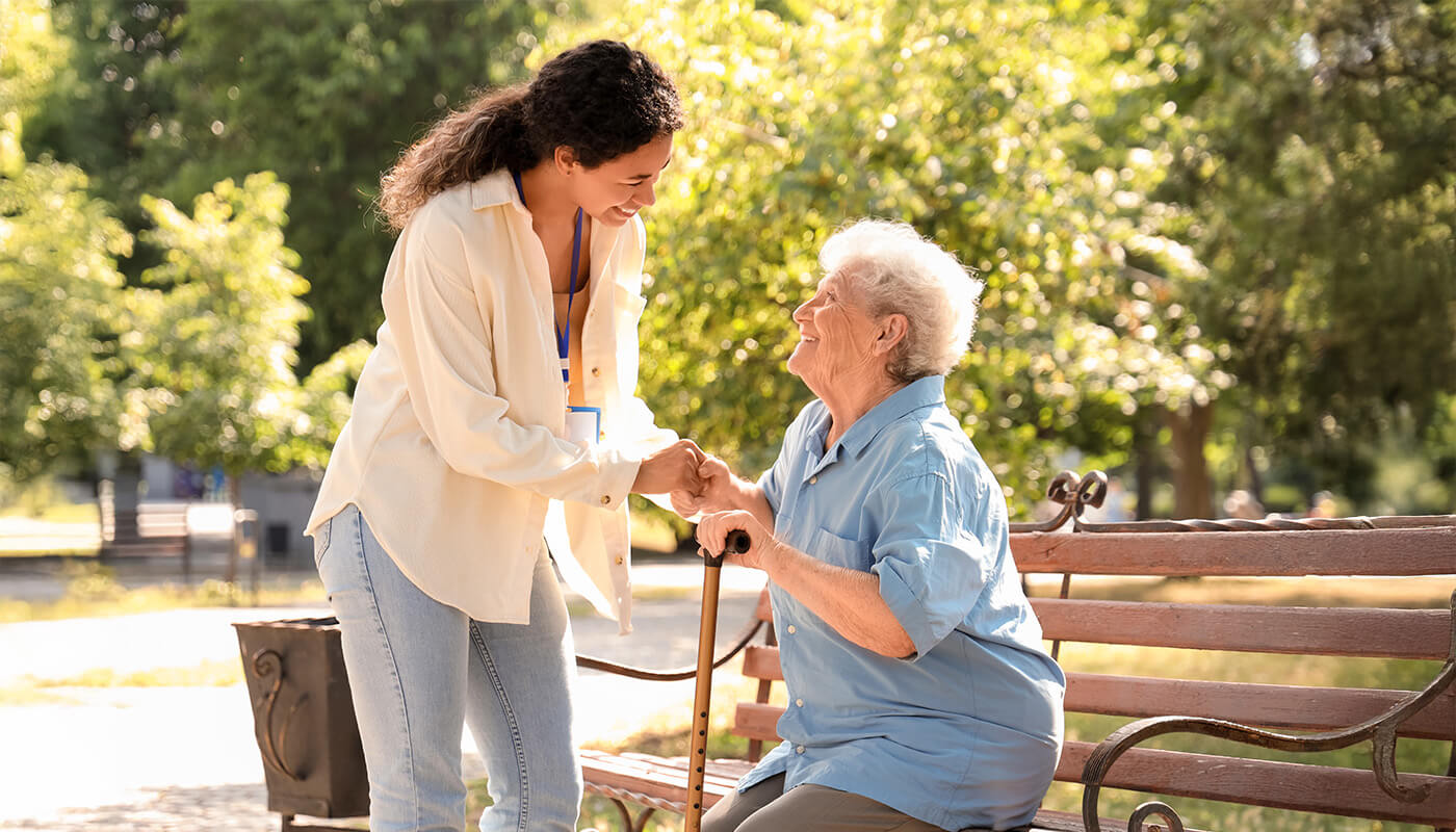 Senior woman sits on an outdoor bench smiling up at younger female caretaker.